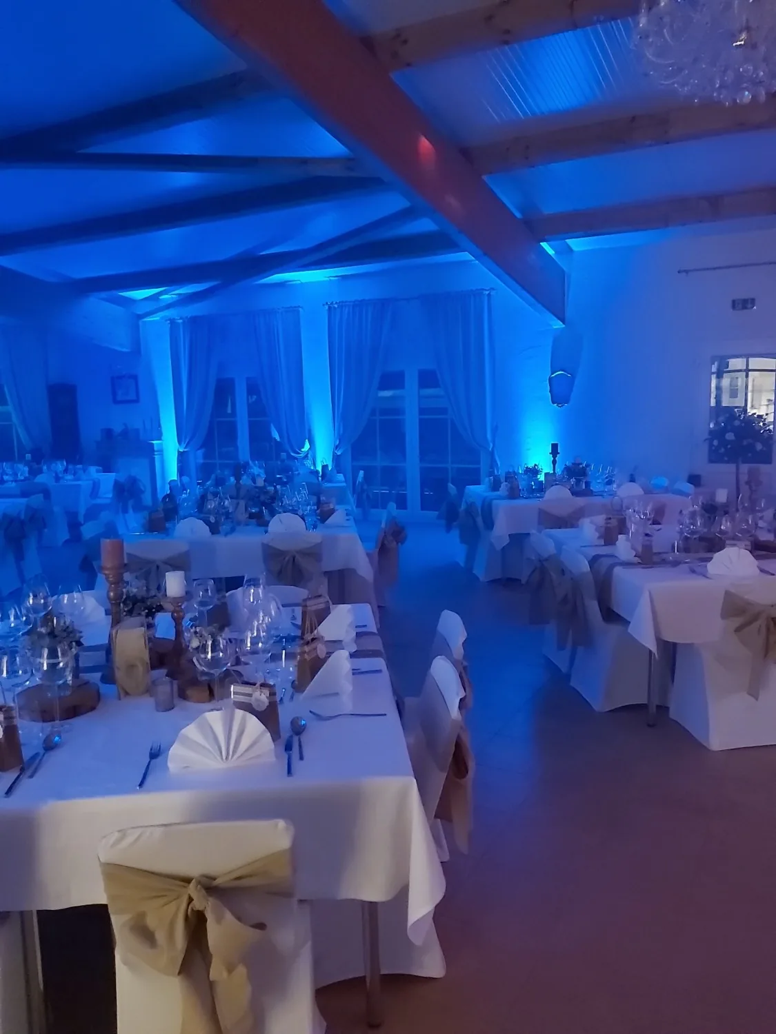 Blau angestrahlter Saal am Abend mit weißen Stuhlhussen, Sackleinen-Schleifen und Kronleuchter unter dem Holzdachstuhl