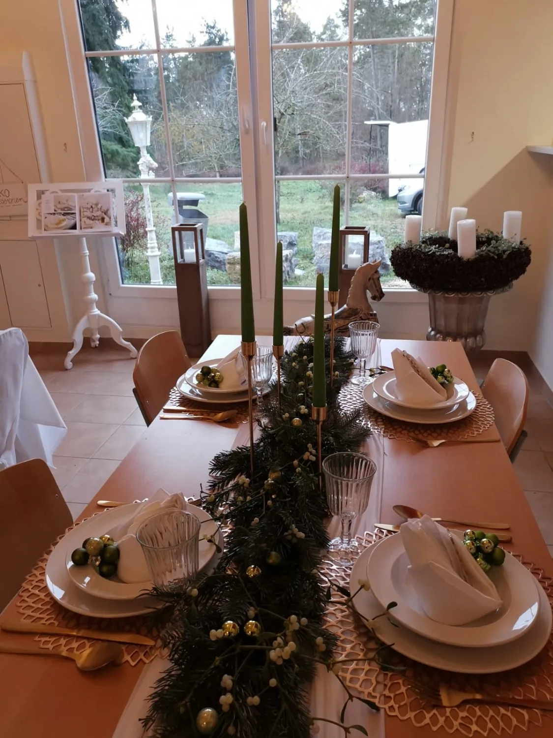 Weihnachtlich gedeckte Tafel mit grünen Kerzen, Tannengirlande und Adventskranz, im Fenster der Blick in den Garten