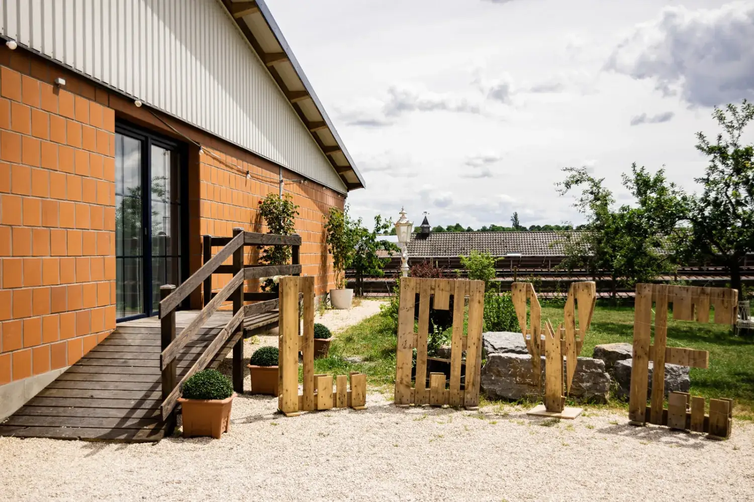 Holzterrasse mit Rampe am Saaleingang, davor Buchsbaumkübel und große Buchstaben aus Paletten auf dem Kiesplatz