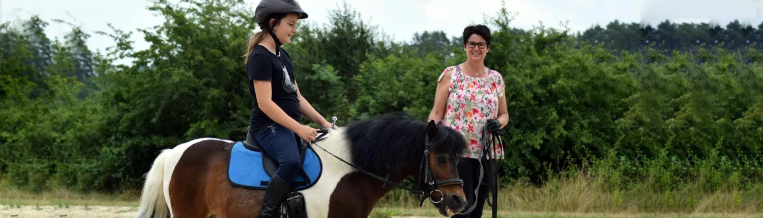 Kind mit Reithelm sitzt auf einem Scheckpony, das die Reitlehrerin über den Sandplatz führt