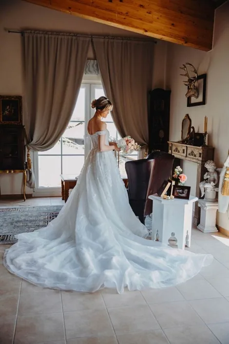 Braut im weißen Kleid mit langer Schleppe am Fenster