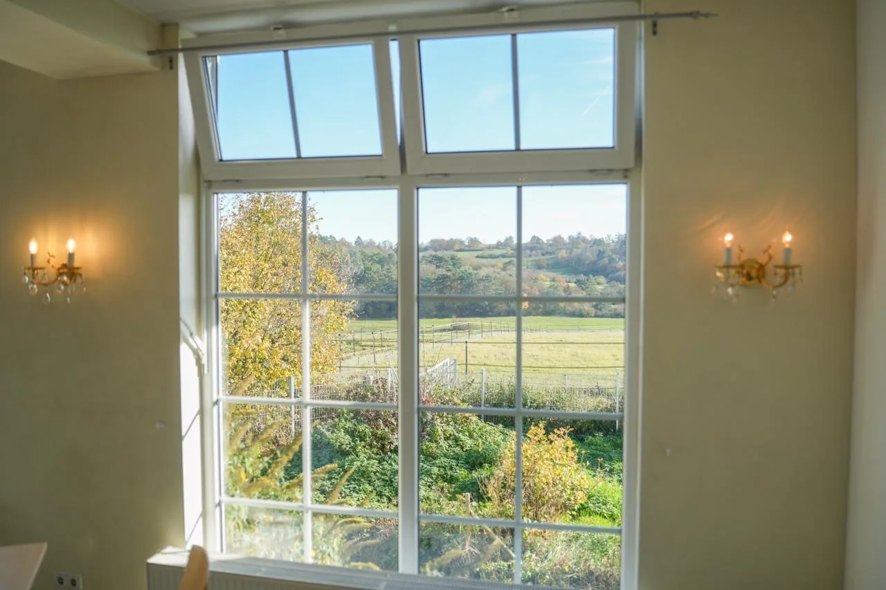 Hohes Sprossenfenster mit Blick über Wiesen und herbstlich gefärbte Bäume, daneben zwei Wandleuchter
