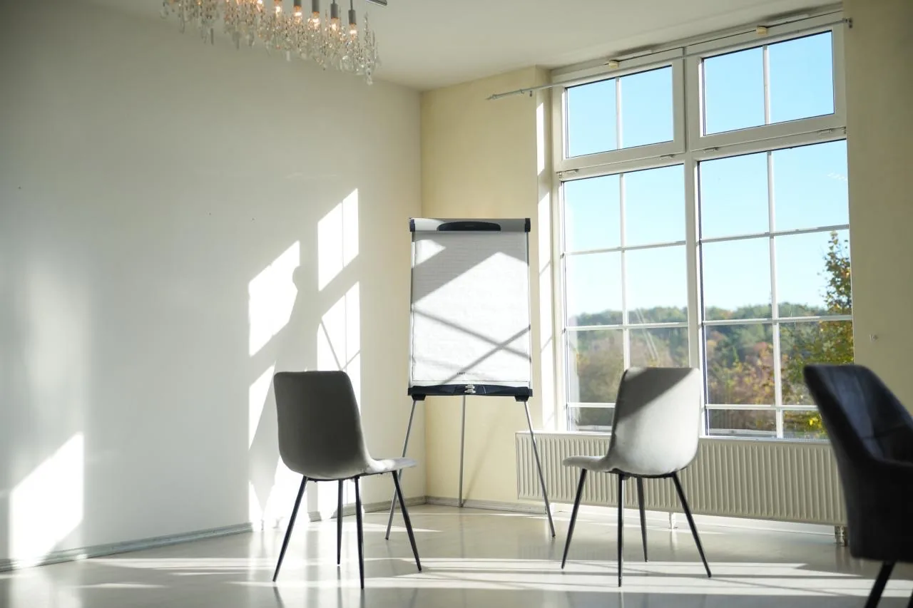Flipchart mit zwei Stühlen in der Salle de Pluvinel, darüber ein Kronleuchter, dahinter das Fenster mit Blick ins Grüne