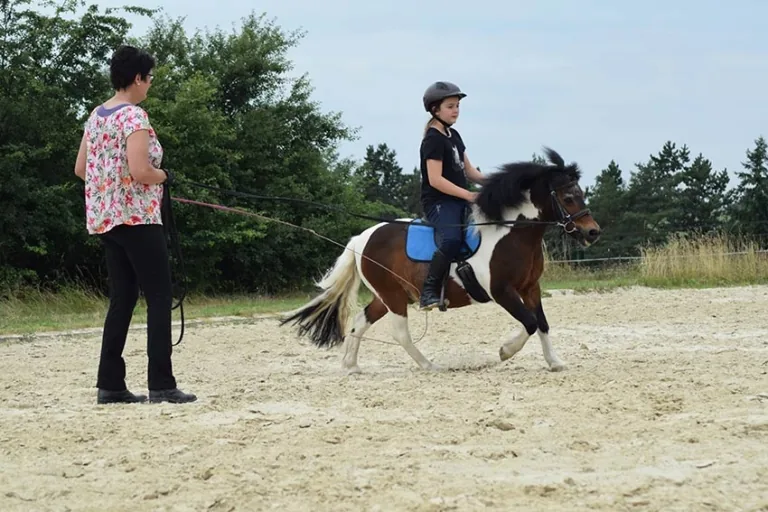 Kind mit Reithelm galoppiert auf einem Scheckpony an der Longe über den Sandplatz, daneben die Reitlehrerin