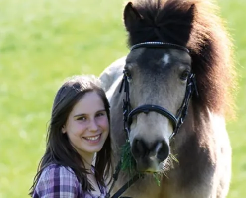 Junge Reiterin steht Kopf an Kopf neben einem falben Pony mit Trense auf der Wiese
