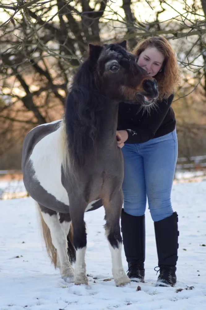 Katharina steht im Schnee neben einem gescheckten Pony und lehnt sich lächelnd an seinen Hals