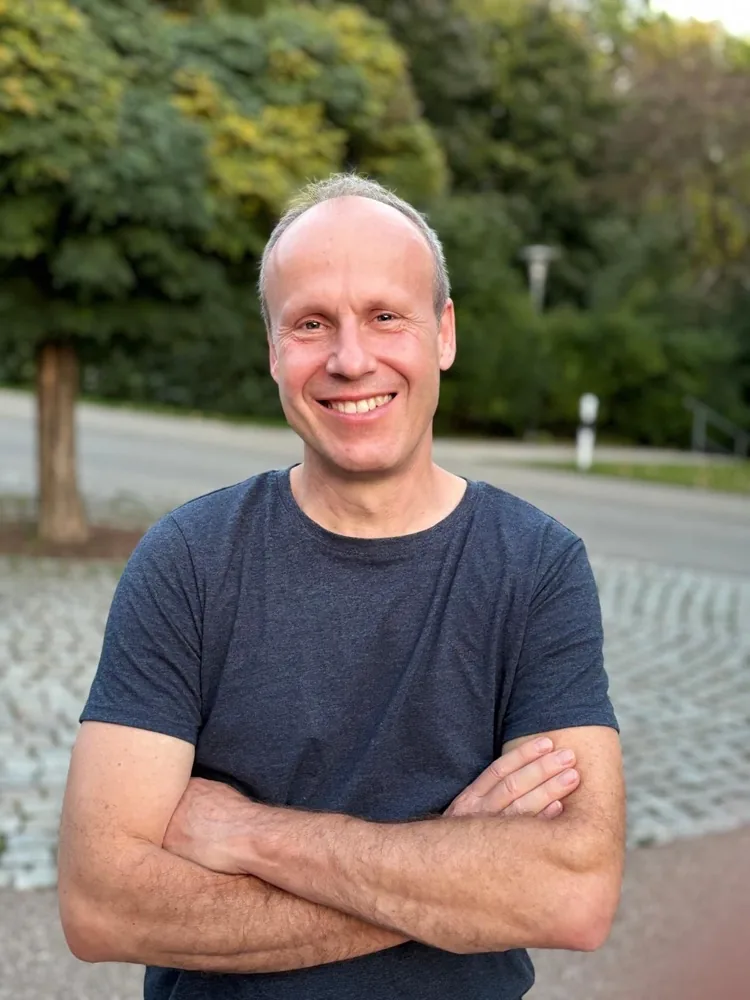 Tom steht mit verschränkten Armen im Freien und lächelt in die Kamera
